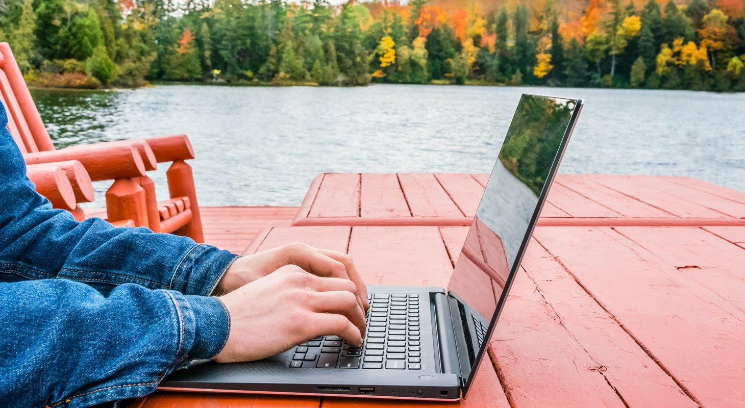 A person working on a laptop outside on a orange table with a view of a lake.